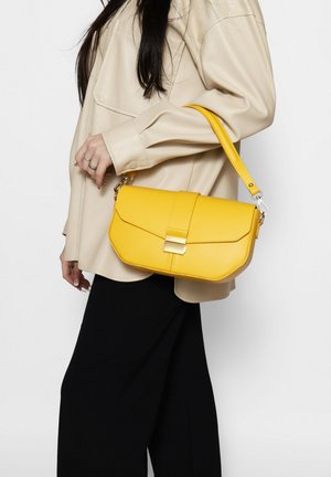 Donna che indossa una camicia beige e pantaloni neri, porta una borsa a mano in pelle gialla con chiusura dorata e tracolla sulla spalla.