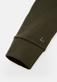 Ciemnozielona bawełniana koszula z długim rękawem, mankiet z ribbowanym wzorem i małym szarym logo przy otworze rękawa. Czysty, minimalistyczny design.