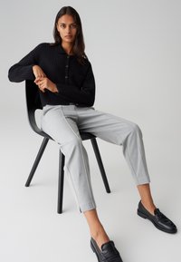 Schwarzer gerippter Strickpullover mit Tasche, hellgraue schmal geschnittene Hose mit Seitennähten und schwarze Loafer, sitzen auf einem schwarzen Stuhl.
