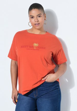 Frau mit kurzen Haaren, die goldene Creolen, ein oranges T-Shirt mit Palme-Stickerei und blaue Jeans trägt, posiert mit einer Hand in der Tasche.