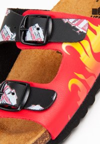 Sandales rouges avec des sangles noires et un motif de feu, comportant des boucles et une semelle en liège, mettant en valeur une surface texturée et des accents colorés.