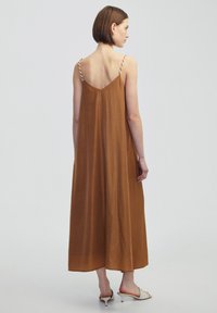 Vestido maxi marrón con tirantes finos y espalda en forma de V. La tela es ligera y de textura suave, con un silueta relajada.