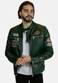 Veste de moto en cuir vert avec un design de manches matelassées, plusieurs patchs, poches zippées et une silhouette ajustée.