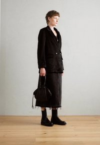 Blazer noir ajusté avec un col cranté, associé à une longue jupe en denim noire avec un ourlet frangé. Le mannequin tient un petit sac à main noir et porte des bottes noires.