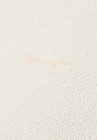 Nahaufnahme eines strukturierten, cremefarbenen Strickstoffs mit dezent eingesticktem "GANT"-Logo in passendem Faden.
