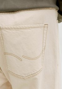 Κοντινή λήψη της πίσω τσέπης μπεζ παντελονιού με καφετιά ραφές και διακριτικές καμπυλωτές διακοσμητικές ραφές.