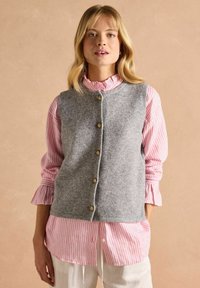 Grauer Wollwesten mit Rundhalsausschnitt, goldenen Knöpfen, über einem gestreiften Langarmshirt in Rosa und Weiß mit Rüschensäumen getragen.
