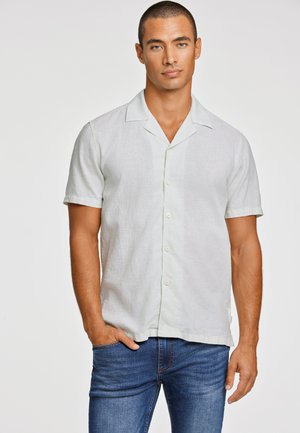 Chemise vert clair à manches courtes en lin avec un col de type camp, dotée d'un devant boutonné et d'une texture subtile. Portée avec un jean bleu.