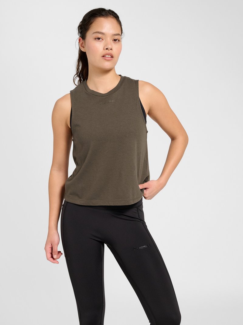Young woman with long dark hair in a ponytail wearing a sleeveless olive workout top and black athletic leggings against a plain light background.