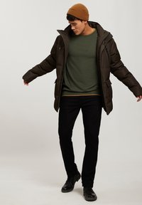 Kurtka puchowa w ciemnozielonym kolorze z kieszeniami zapinanymi na zamek, noszona na ciemnozielonym swetrze. Czarna para dżinsów i czarne buty dopełniają strój. Brązowa czapka akcentująca.