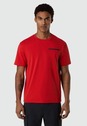 Uomo che indossa una maglietta rossa a maniche corte con un piccolo logo nero sul petto, in piedi davanti a uno sfondo grigio chiaro.