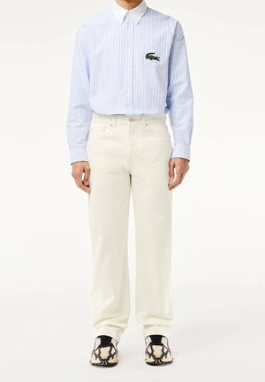 Mann trägt hellblau-weiß gestreiftes Langarmhemd mit Krokodil-Logo, weiße Hose und schwarz-beige gemusterte Sneakers.