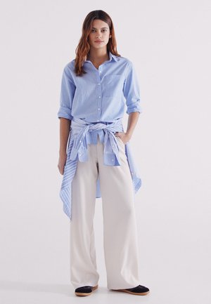 Blau gestreiftes Hemd mit aufgerollten Ärmeln, am Taillenbund gebunden über einer hellbeigen, weiten Hose und schwarzen Schuhen. Sauberes, modernes Design.