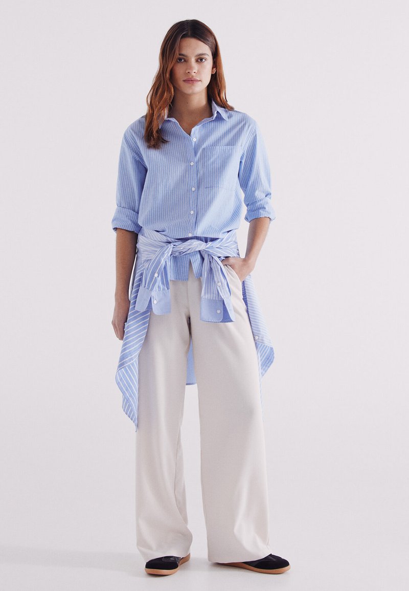 Blue striped button-up shirt with rolled sleeves, tied at the waist over light beige wide-leg pants and black shoes. Clean, modern design.