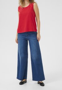 Top sin mangas rojo combinado con jeans de mezclilla azul de pierna ancha. El top tiene un escote redondo, mientras que los jeans presentan un corte y acabado clásicos.