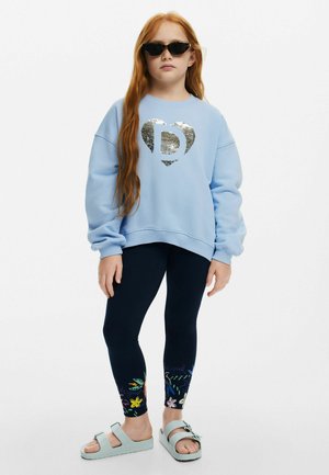 Hellblaues, übergroßes Sweatshirt mit einem silbernen, paillettenbesetzten Herzensdesign, kombiniert mit marineblauen Leggings mit Blumenmustern und hellen Sandalen.