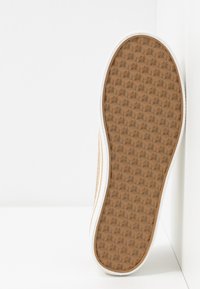 Semelle d'une basket beige avec bordure blanche, présentant un motif géométrique pour l'adhérence, montrée debout sur un fond blanc.