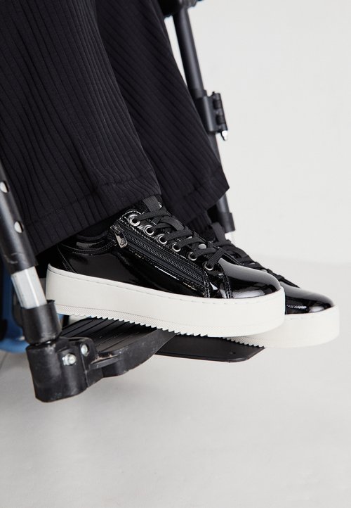 Zapatillas de charol negro con superficie texturizada, que cuentan con una suela de plataforma blanca, cierre de cordones en la parte frontal y detalle de cremallera en el lateral.