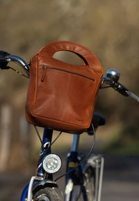 Brun läderhandväska med en strukturerad design, rund topphandtag och framficka med dragkedja, placerad på en cykelhandtag.