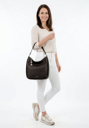 Lächelnde Frau, die eine beige Bluse und weiße Hose trägt, hält eine schwarze Lederhandtasche mit silbernen Anhängern und steht vor weißem Hintergrund.