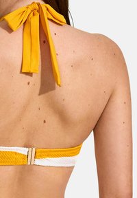 Haut de bikini jaune avec une sangle texturée et un accent blanc, noué avec un nœud assorti à l'arrière, mettant en valeur un teint de peau lisse.