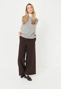 Grauer ärmelloser Strickpullover mit einem Rippbund, kombiniert mit hoch taillierten, weit geschnittenen braunen Hosen und dunklen Loafers. Glatte Texturen.