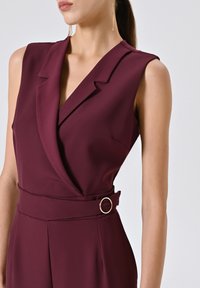 Tuta senza maniche color bordeaux con scollatura a V, vita arricchita da una cintura con un accento circolare dorato e tessuto liscio e texturizzato.