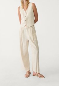 Beige linnen vest met vijf houten knopen, gecombineerd met beige geplooide broek met losse pasvorm en lichte sandalen met gekruiste bandjes.