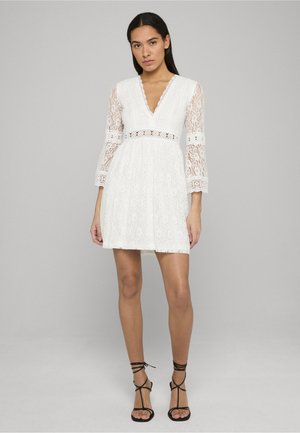 Hvid blondekjole med V-hals og klokkeærmer, har en cirkulær udskæring ved taljen. Kombineret med sorte sandaler med stropper.