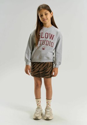 Pige med langt brunt hår iført grå sweatshirt med teksten "SLOW STUDIO", brun zebra-mønstret nederdel, sokker med hjerteprint og beige sneakers.