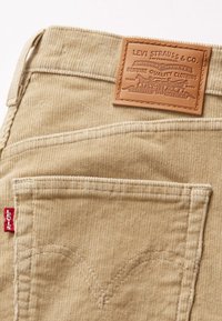 Tejido de pana beige con líneas verticales, que presenta una etiqueta de cuero grabada con texto y una etiqueta roja de Levi's en el bolsillo lateral.