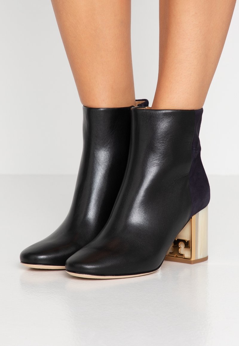 Bottines en cuir noir avec talons carrés dorés portées sur des jambes nues sur fond blanc uni.