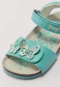 Turkis sandaler med en blank finish, dekorert med sølvglitrende sommerfugldetaljer og blomsterformer på sålen. Justerbar reim.