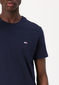 Persoană cu pielea închisă la culoare care poartă un tricou simplu bleumarin cu mânecă scurtă, cu un mic logo dreptunghiular roșu, alb și albastru pe piept.