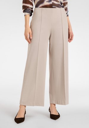 Beżowe spodnie szerokie wykonane z gładkiego, elastycznego materiału, z fałdami z przodu i wysokim stanem. W zestawie z ciemnymi, fakturowanymi butami.
