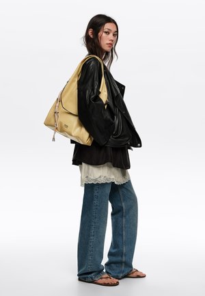 Mujer con chaqueta de cuero negra, camisetas superpuestas, vaqueros anchos azules, sandalias y un bolso grande beige de hombro con detalles de cordón.