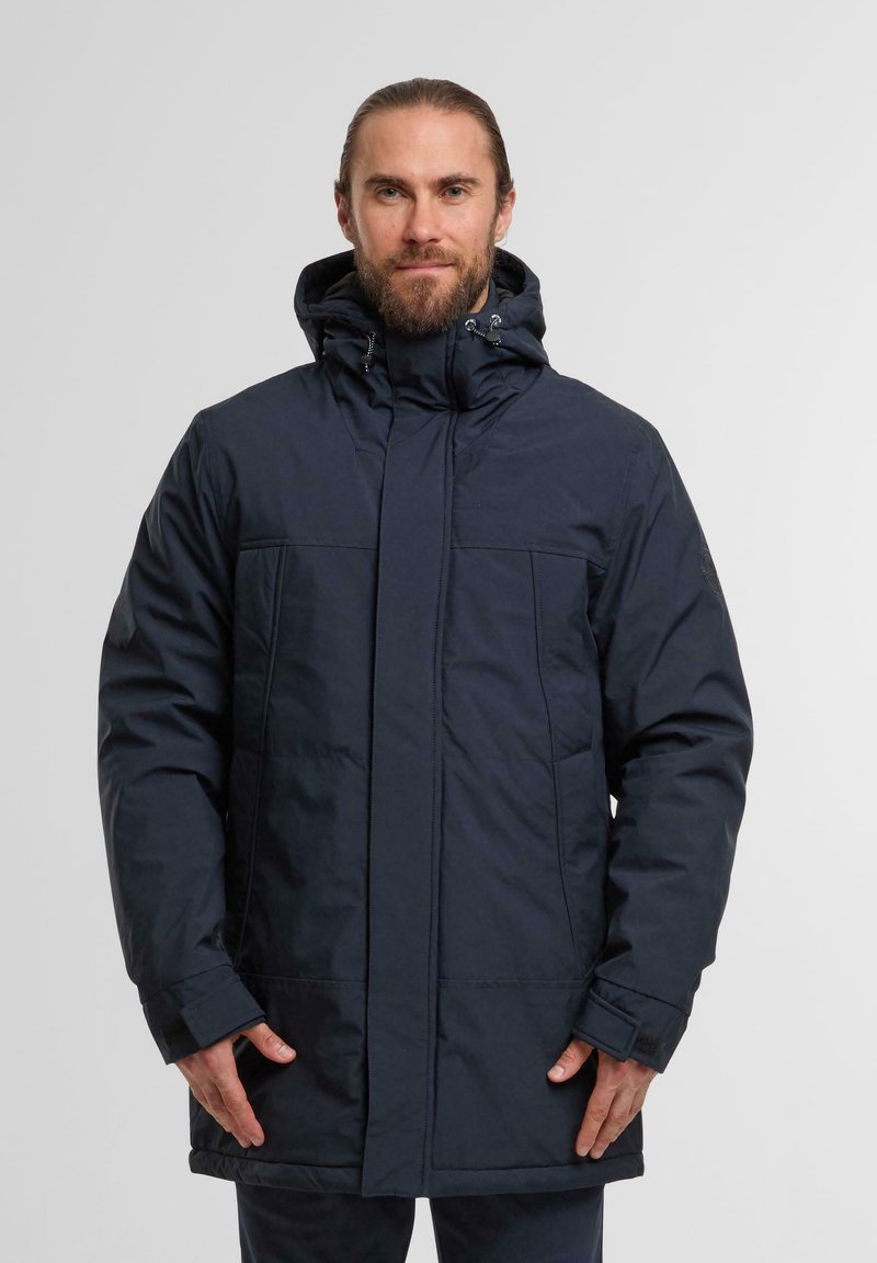 Giacca isolante blu navy con colletto alto, chiusura con zip e bottoni a pressione, e due tasche frontali. Caratterizzata da un cappuccio con cordino regolabile e polsini regolabili.