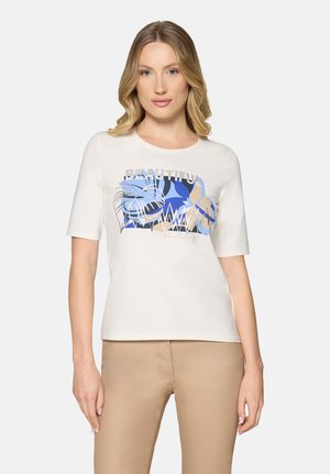 Kobieta o blond włosach, ubrana w biały krótki rękaw z abstrakcyjnym wzorem liści w kolorach niebieskim i beżowym oraz beżowe spodnie, stojąca na jednolitym tle.