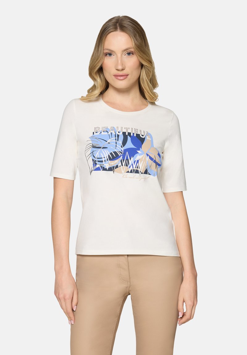 Femme aux cheveux blonds portant un haut blanc à manches courtes avec un motif abstrait de feuilles bleu et beige, et un pantalon beige, debout devant un fond uni.
