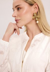 Biała bomberka o fakturowanej powierzchni z zapięciem na zamek, w połączeniu z dużymi złotymi kwiatowymi kolczykami akcentowanymi kremową perełką.