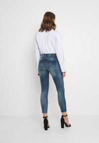 Jeans ajustados en denim azul deslavado con cintura alta, bolsillos traseros y dobladillos deshilachados, combinados con una camisa de botones azul clara y tacones negros.