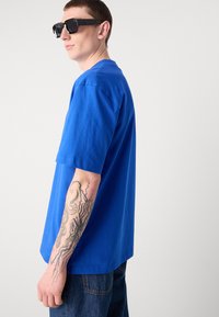 Kongsblå bomullst-skjorte med korte ermer, avslappet passform og rund hals, kombinert med mørke jeans og svarte rektangulære solbriller.
