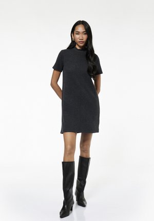 Vrouw die een donkergrijze jurk met korte mouwen en kniehoge zwarte leren laarzen draagt, staat met haar handen achter haar rug op een witte achtergrond.