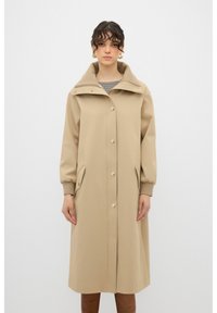 Beige trenchcoat met een grote kraag, voor knopen en zijzakken. De mouwen zijn lichtjes pof, met ribgebreide manchetten voor extra comfort.