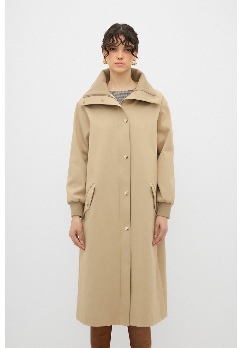 Trench de color beige con un gran cuello, botones en el frente y bolsillos laterales. Las mangas son ligeramente abullonadas y cuentan con puños acanalados para mayor comodidad.