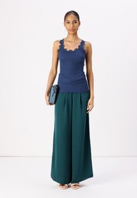 Blauwe geribbelde top met kantafwerking, gecombineerd met groene wijde broekspijpen. Model houdt een zwarte leren clutch vast en draagt naaktsandalen. Eenvoudige achtergrond.