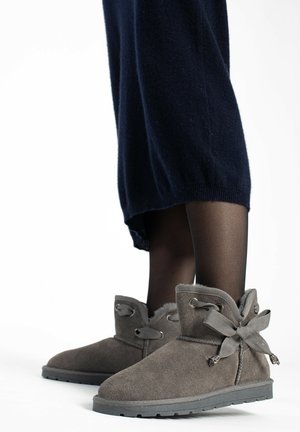 Botines de ante gris con un interior suave, acentos de cinta y suela de goma. Llevados con medias negras y una prenda de punto oscura.