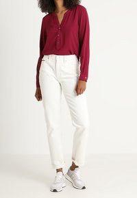 Bordeaux knoopblouse met een v-hals, gecombineerd met witte rechte jeans en lichtgrijze sneakers met zwarte accenten.