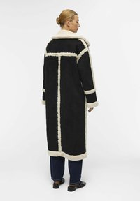 Schwarzer langer Mantel mit Schaffellbesatz und Kragen. Verfügt über vertikale und horizontale Paneele. Getragen über dunklen Jeans und Schuhen.