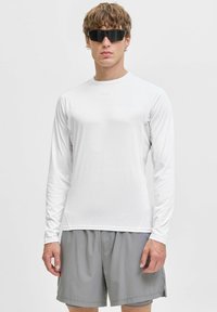 Vit långärmad skjorta med en slät textur, parat med grå shorts. Modellen bär svarta solglasögon. Enkelt, atletiskt mönster, inga mönster.